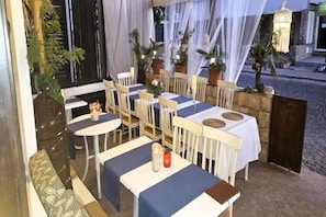 Un área de comedor al aire libre con mesas y sillas, algunas con manteles, y plantas en macetas.