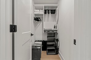 A narrow hallway with a closet containing hanging clothes, shelves with folded items, and a small table with a kettle.