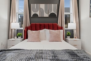 A modern bedroom with a red upholstered headboard, two bedside tables with lamps, and a large abstract artwork above the bed. - Designer Condo w Parking | Zoo, Museums • Center City (Philadelphia)