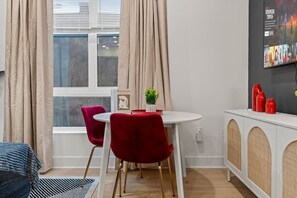 A modern living room with a dining area, featuring a white round table, red velvet chairs, a small potted plant, and a bookshelf. - Designer Condo w Parking | Zoo, Museums • Center City (Philadelphia)