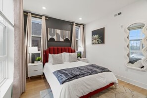 A modern bedroom with a red upholstered headboard, a large bed with white bedding, a bedside table with a lamp, and a wall-mounted mirror.