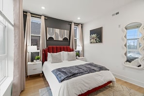 A modern bedroom with a red upholstered headboard, a large bed with white bedding, a bedside table with a lamp, and a wall-mounted mirror. - Bright Boutique Condo | Parking | Downtown•Univ City (Philadelphia)