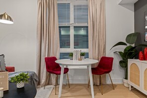 Ein Zimmer mit einem runden weißen Tisch, zwei roten Stühlen, einem Bücherregal und einem Fenster mit Vorhängen.