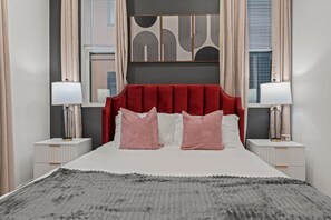 Une chambre à coucher avec une tête de lit rembourrée de tissu rouge, deux tables de chevet avec des lampes, un lit avec des oreillers roses et un tapis gris à texture intéressante.