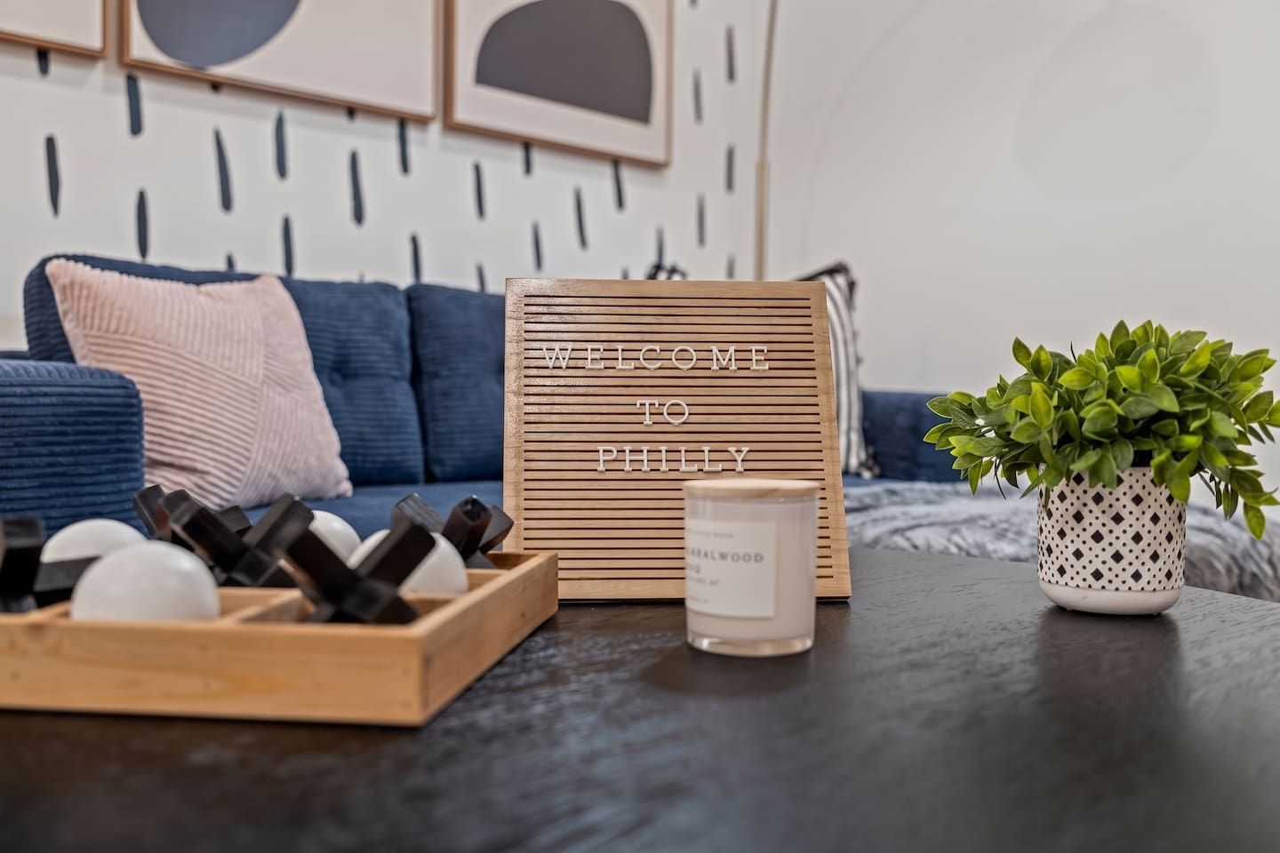 Une enseigne en bois portant l’inscription « BIENVENUE À PHILADELPHIE », une bougie, une plante en pot et un plateau contenant des objets sphériques, le tout posé sur une table.