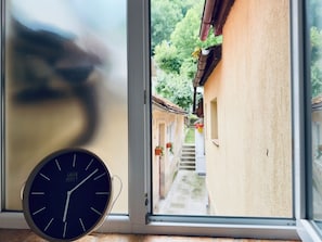 Una habitación con un reloj en la pared y una ventana con vistas a una calle estrecha con edificios y vegetación.