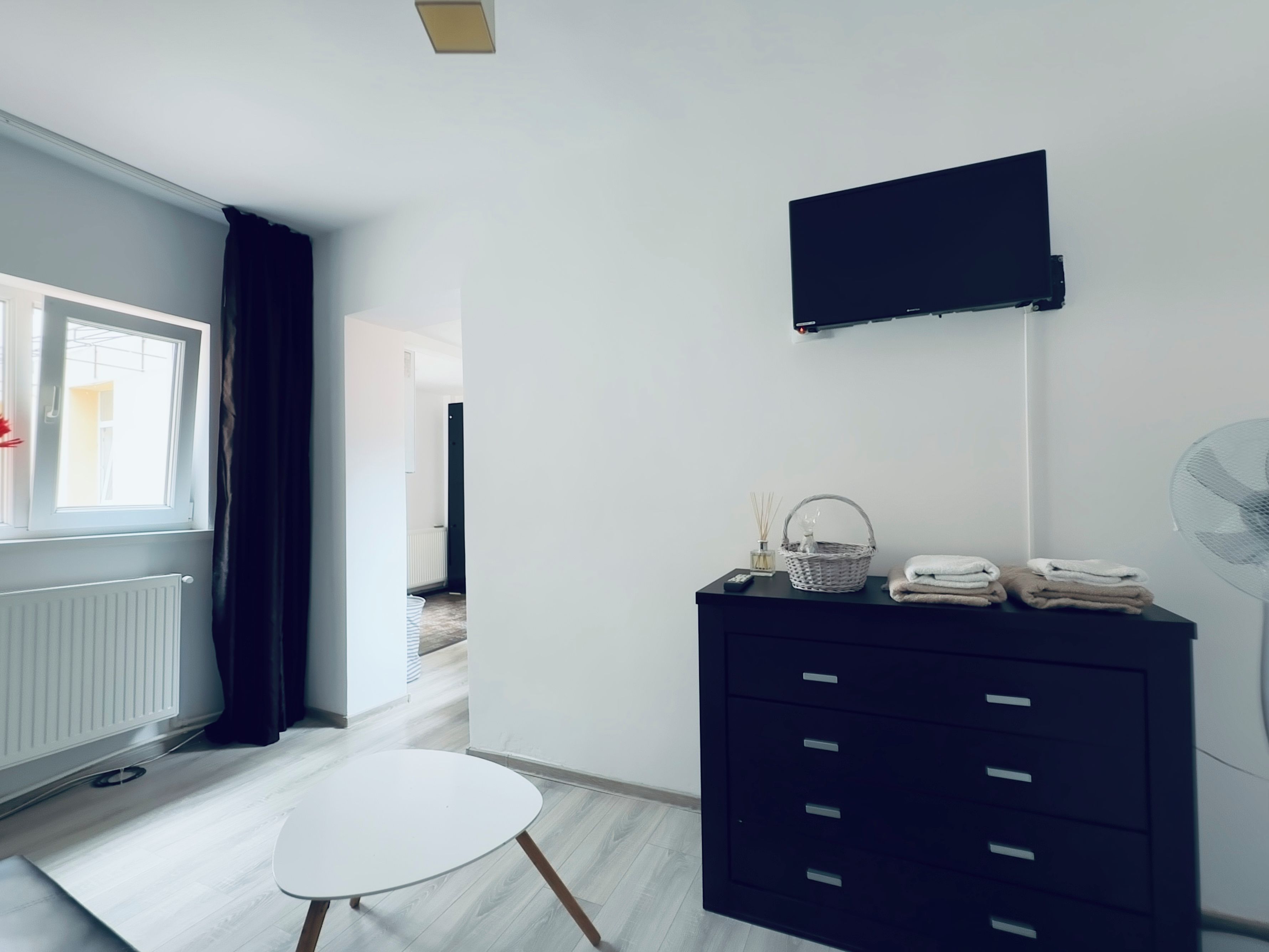 Una habitación moderna con una mesa redonda blanca, un aparador negro con cajones, un televisor montado y una ventana con cortinas.