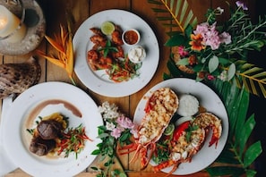 Una mesa con platos de comida, incluyendo mariscos y un postre, rodeada de flores decorativas y follaje.
