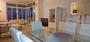A modern living room with a glass dining table, a sofa, a TV, and a view of the city at night. - Apartamento con Espectaculares Vistas (Palma de Mallorca)