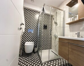 A modern bathroom with a geometric patterned floor, a glass-enclosed shower, a white toilet, and a wooden vanity with a mirror. - Dugi (Istanbul)