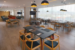 Um restaurante moderno com várias mesas e cadeiras, janelas amplas e iluminação pendente.
