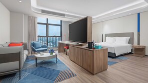 Ein modernes Hotelzimmer mit einem großen Bett, einem Schreibtisch mit Flachbildfernseher, einer Couch und einem Balkon mit Blick auf die Stadt.