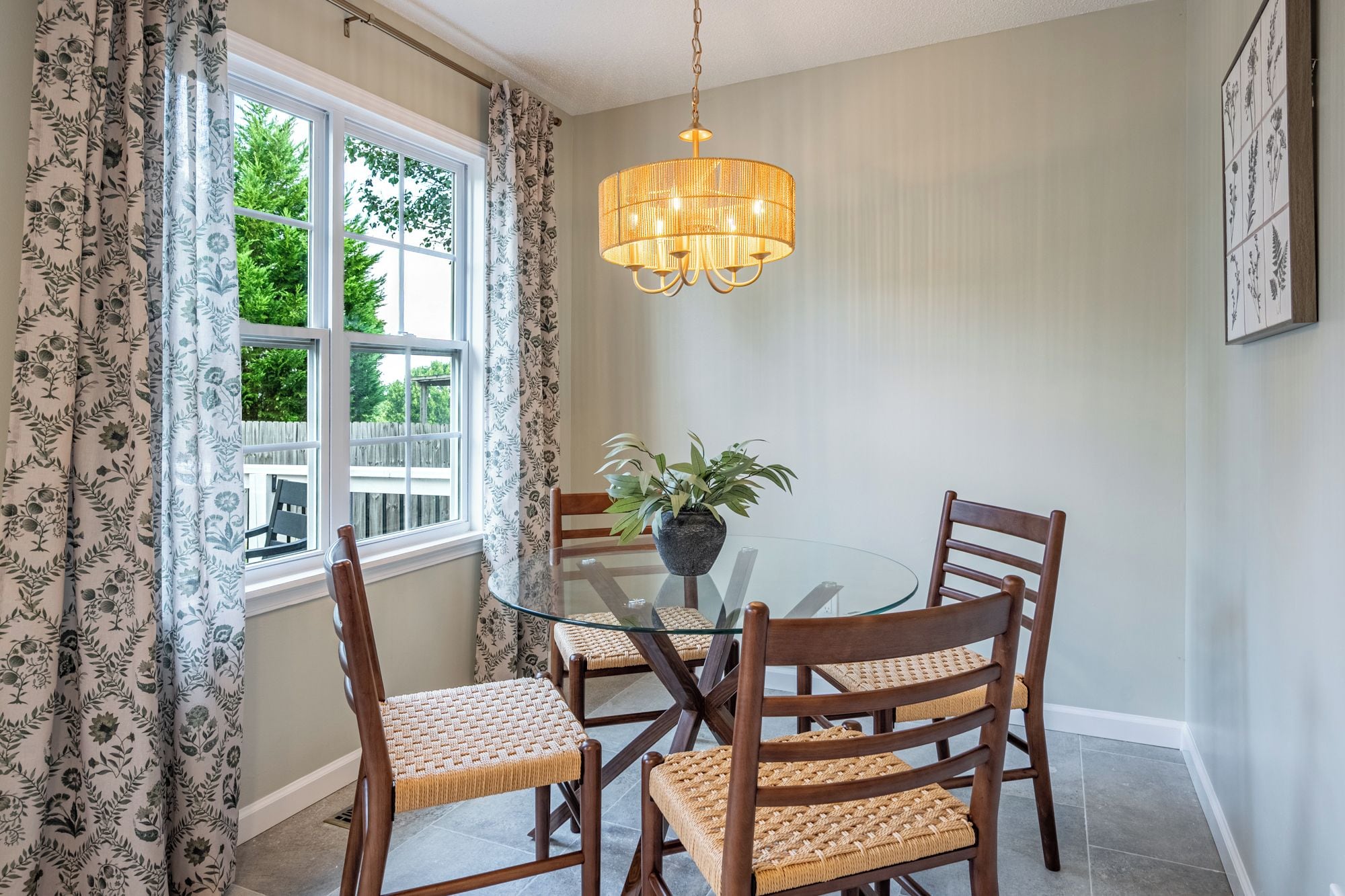 Et spiseområde med et glasbord omgivet af træstole med flettede sæder, en hængende lampe og et vindue med mønstrede gardiner.