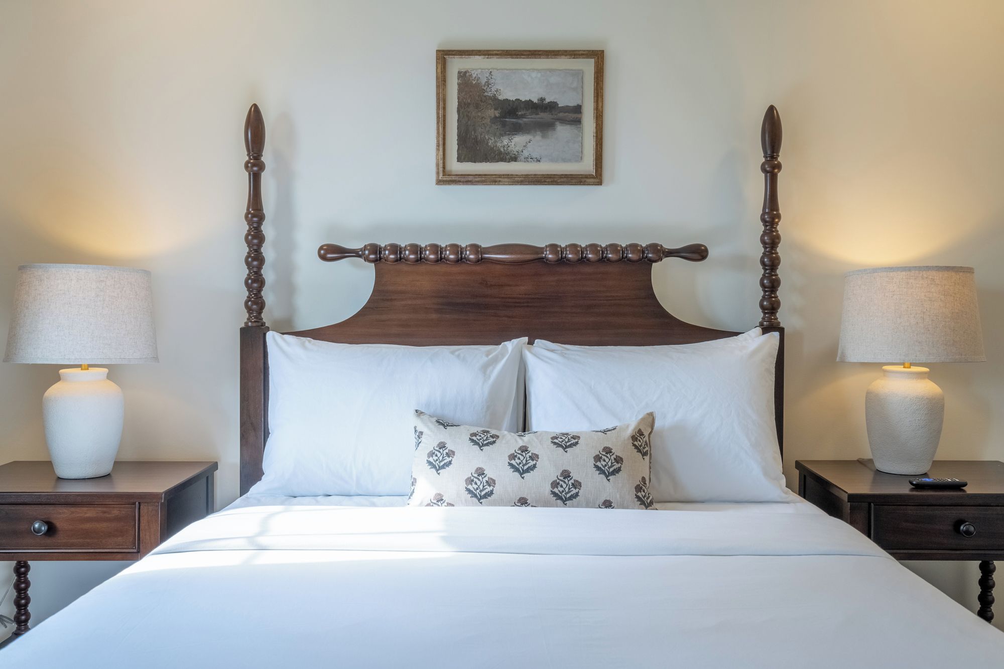 Een hemelbed met witte lakens en een kussen met een patroon, met aan beide zijden nachtkastjes met lampen, in een kamer met een kunstwerk erboven.