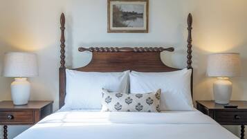 Een hemelbed met witte lakens en een kussen met een patroon, met aan beide zijden nachtkastjes met lampen, in een kamer met een kunstwerk erboven.