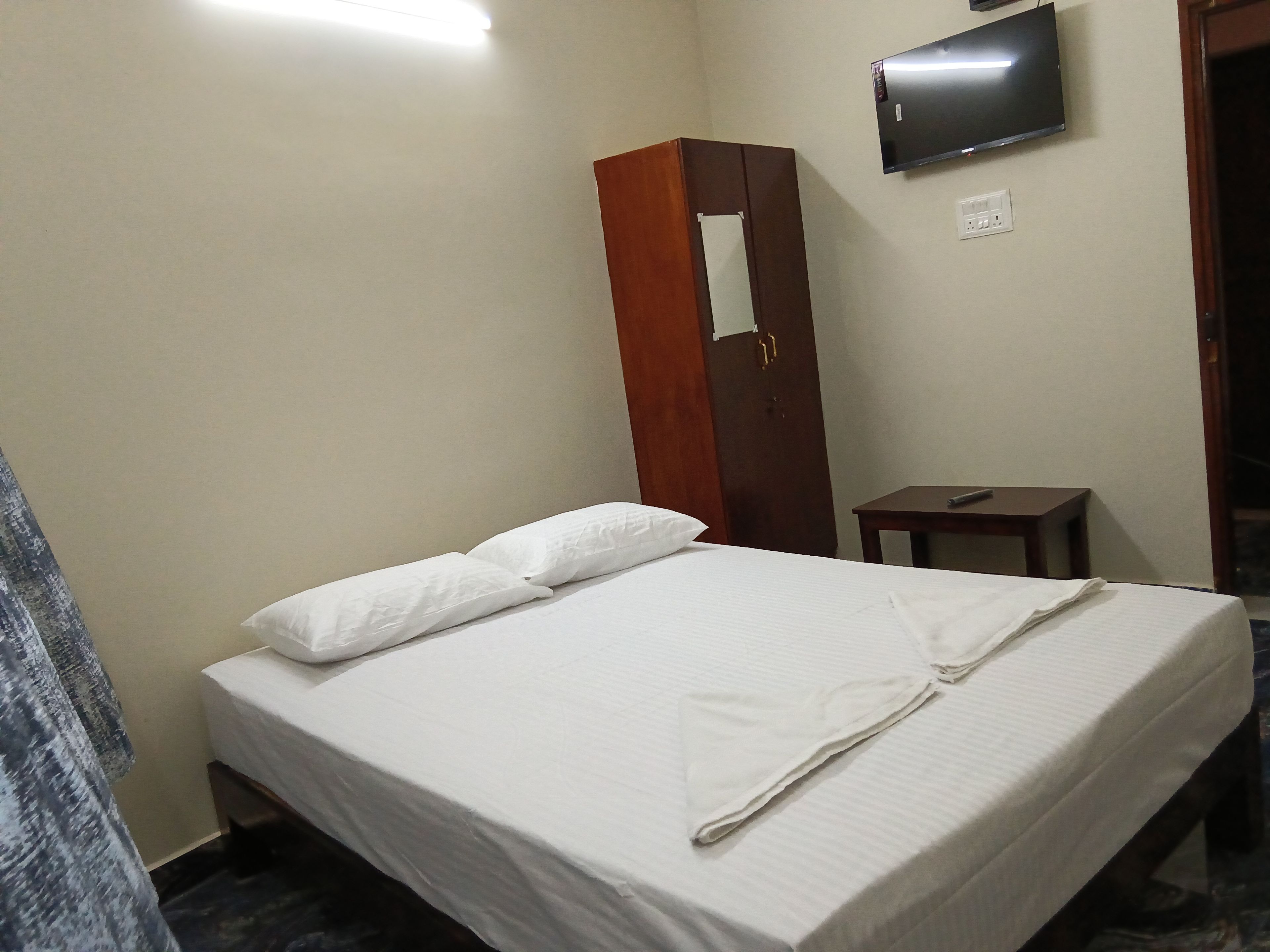 A hotel room with a bed, a wooden wardrobe, a small table, and a television mounted on the wall.