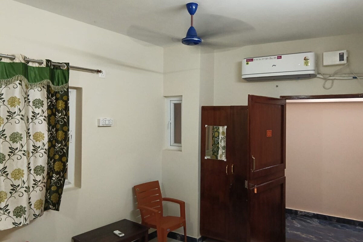 Una habitación con ventilador de techo, aire acondicionado, armario de madera, una silla de plástico y una mesita.