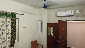 Una habitación con ventilador de techo, aire acondicionado, armario de madera, una silla de plástico y una mesita.