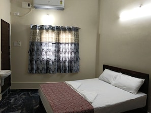 A hotel room with a bed, a window with patterned curtains, an air conditioning unit, and a wall socket.