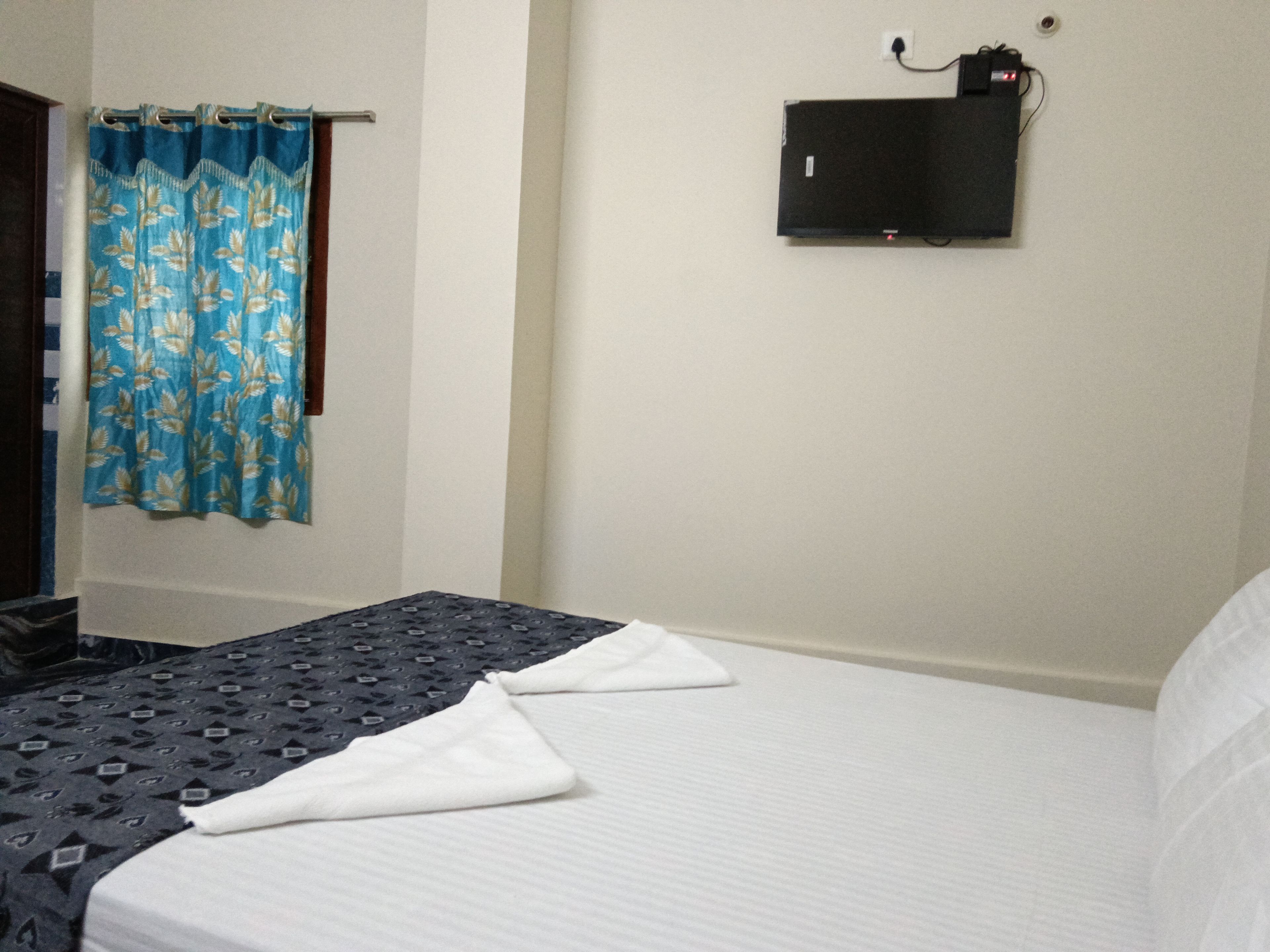 A hotel room with a bed, a flat-screen TV mounted on the wall, and a curtain with a leaf pattern.