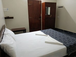 A single bed with a white mattress and a patterned blanket, a wooden wardrobe, a small bedside table, and a wall socket.