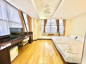 Habitació d'hotel amb un llit, un escriptori amb televisió, un ventilador de sostre i grans finestres amb cortines.