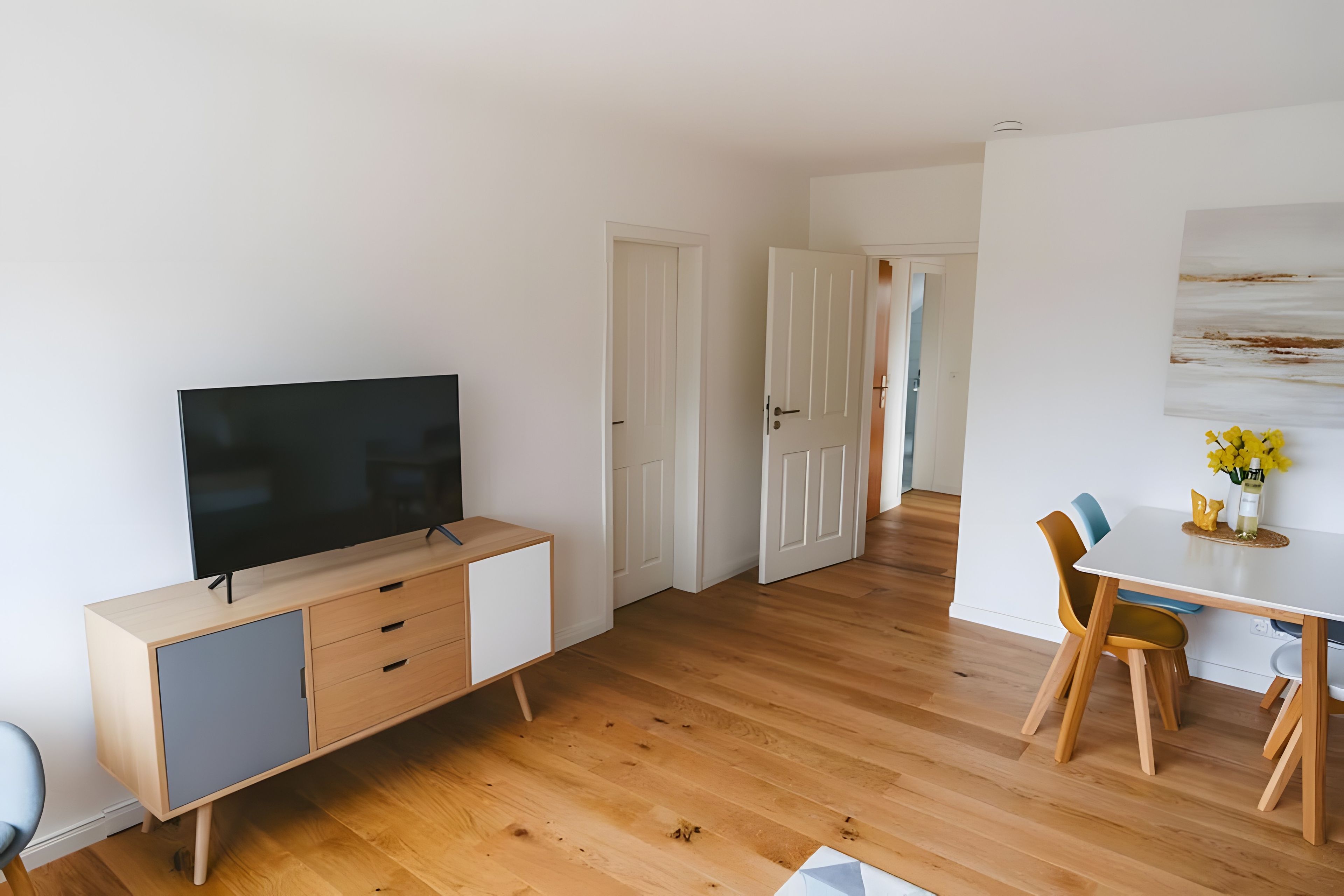 A modern living room with a flat-screen TV, wooden flooring, a dining area with a table and chairs, and a hallway leading to another room.