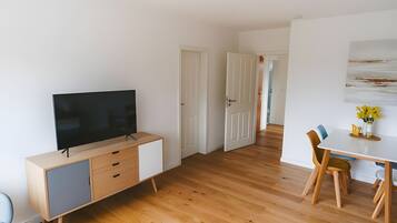 A modern living room with a flat-screen TV, wooden flooring, a dining area with a table and chairs, and a hallway leading to another room.