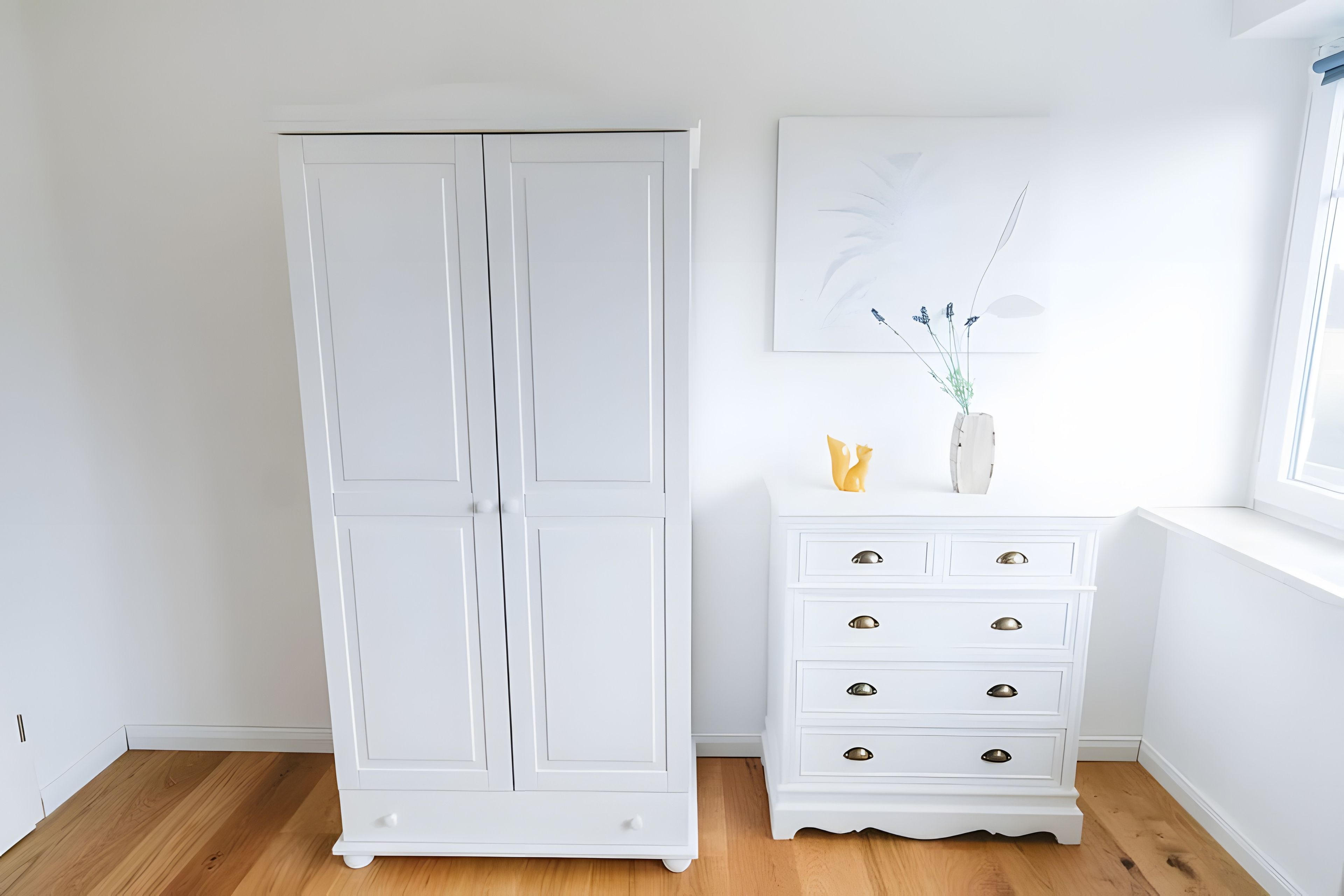 A white wardrobe with a mirror, a white dresser with drawers, and a wall-mounted artwork.