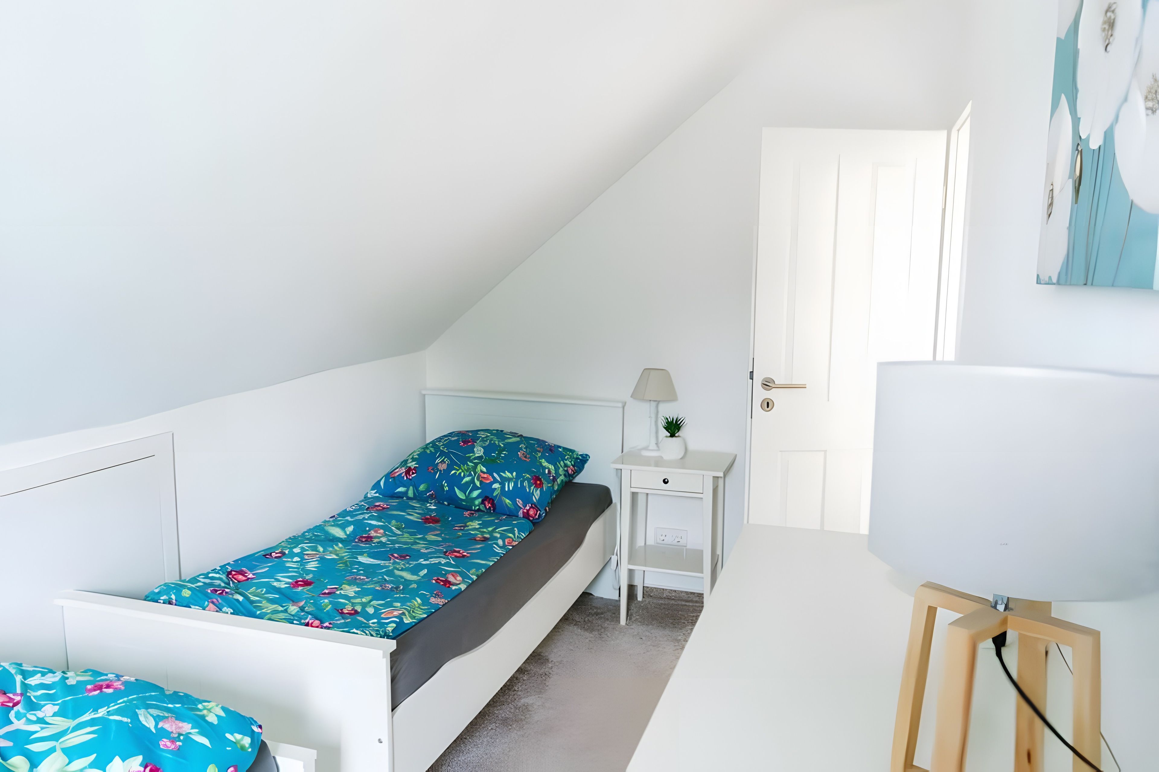 Een kleine kamer met een enkel bed, een schuine kap, een nachtkastje met lamp en een deur naar een andere kamer.