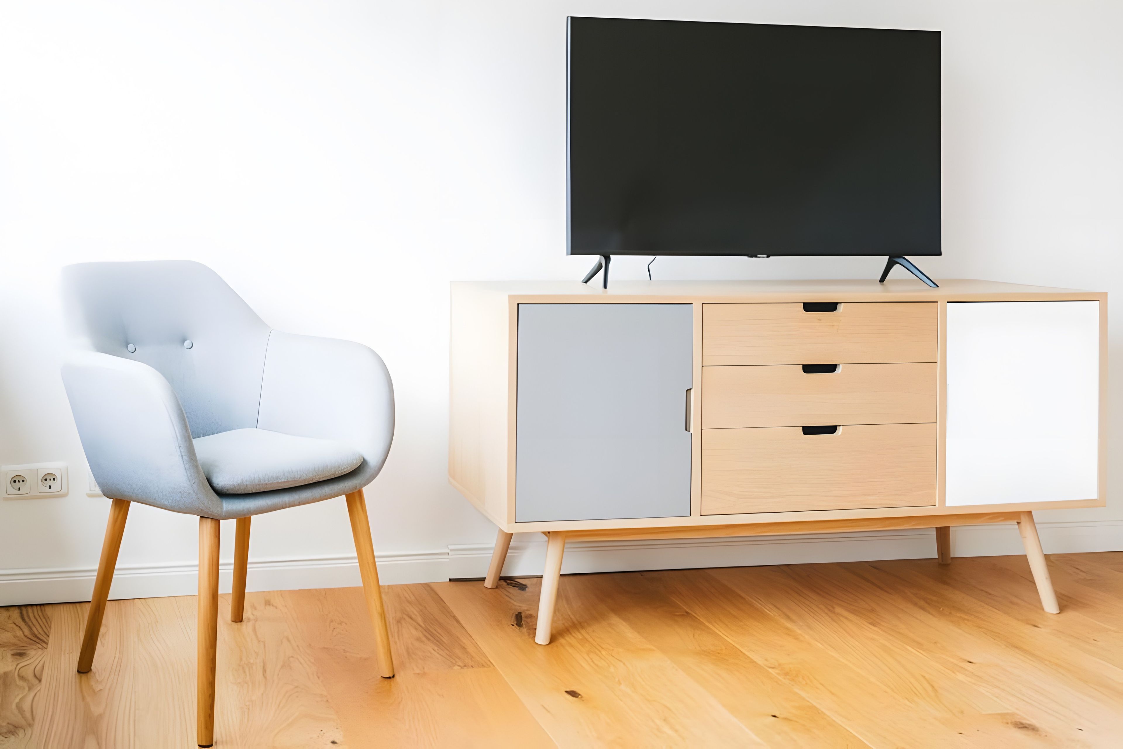 A modern room with a light grey chair, a wooden floor, a light wooden cabinet with drawers, and a black TV on the wall.