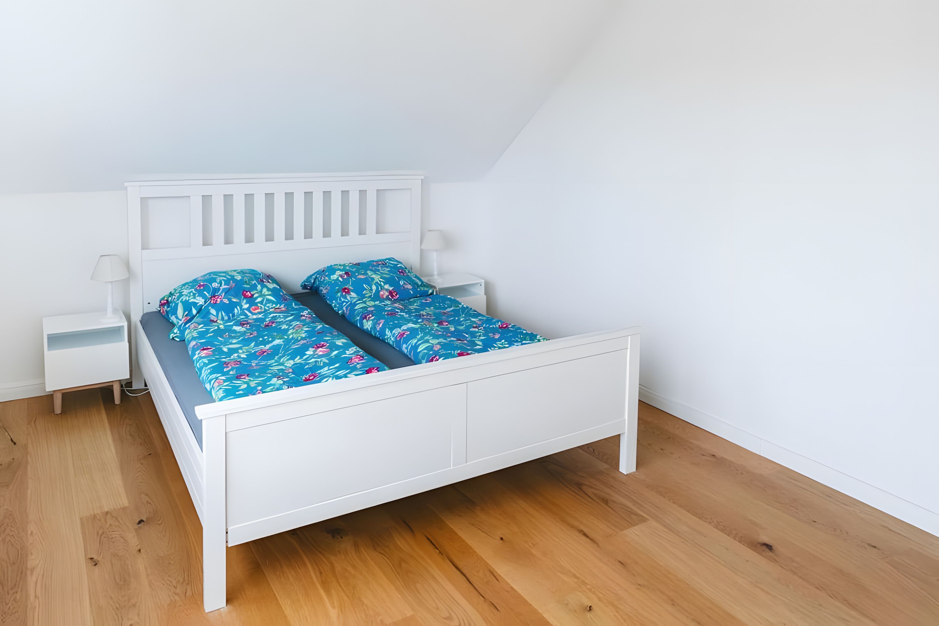 A white bed with a blue floral bedspread and matching pillows, a white nightstand with a lamp, and a wooden floor.