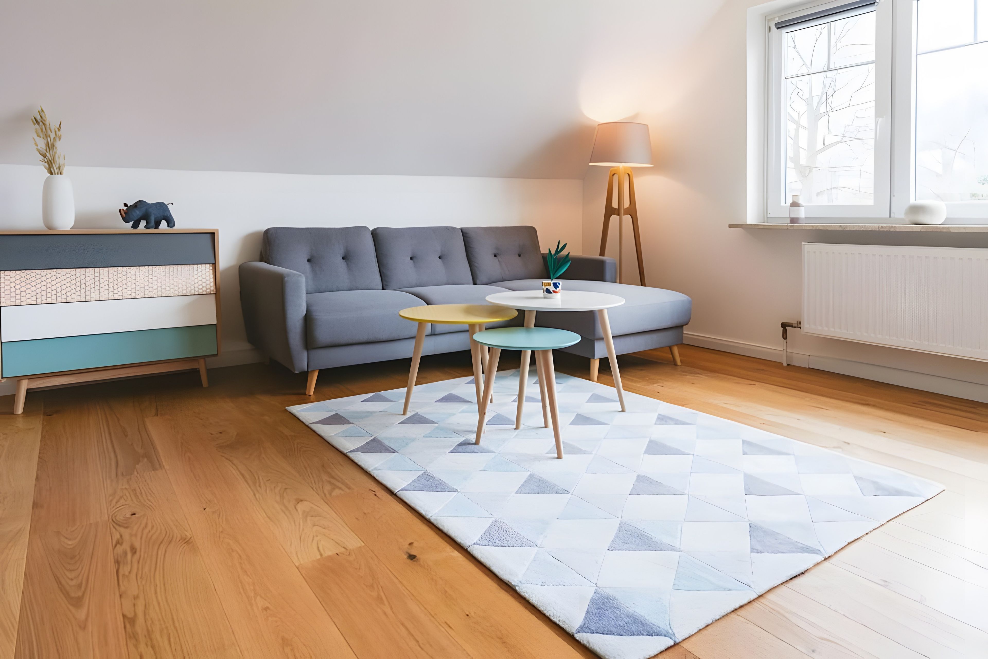 Ein modernes Wohnzimmer mit einem grauen Sofa, Holzboden, einem gemusterten Teppich und einem kleinen runden Tisch mit einer Lampe.