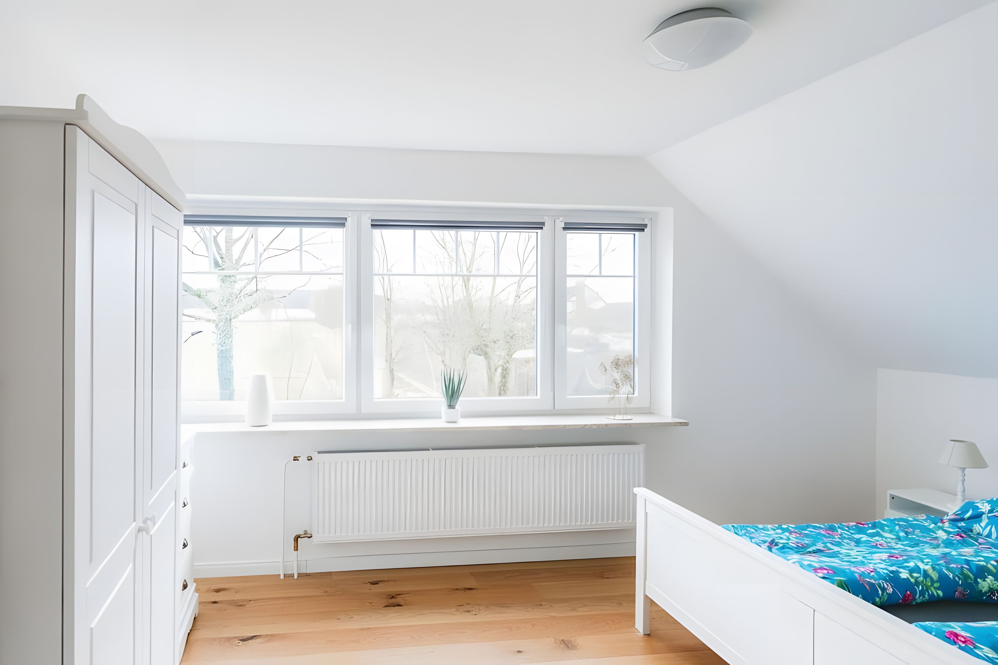 A bedroom with a white radiator, a window with blinds, a bed with a floral bedspread, and a white wardrobe.