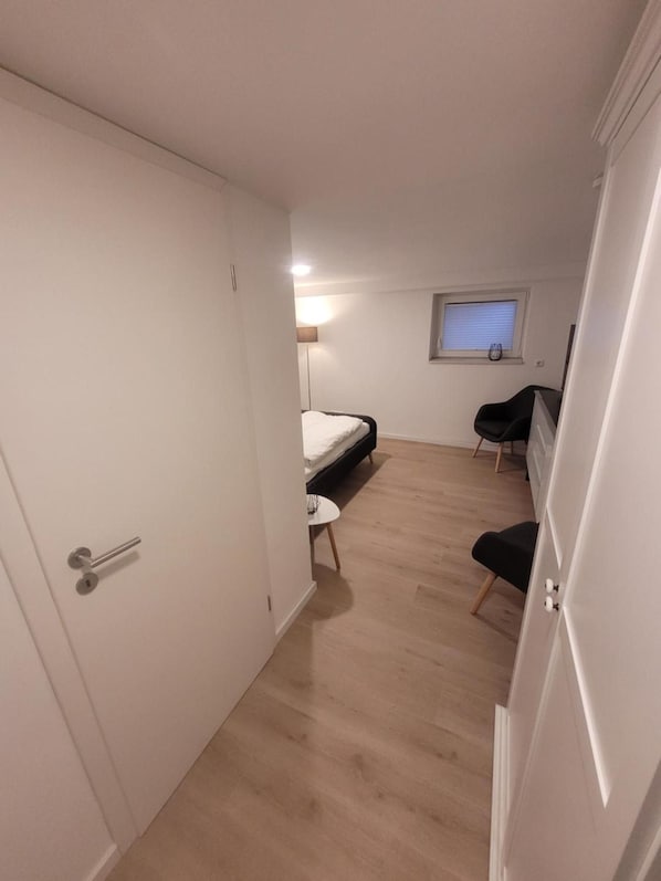 A room with a bed, a desk, and a chair. There is a window with blinds and a lamp on the wall.