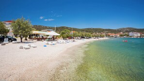 En strand med klart, grunt vatten, en restaurang och en rad solstolar.