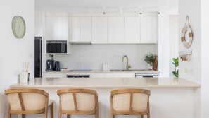 A modern kitchen with white cabinets, a central island with bar stools, a microwave, and a sink with a gold faucet. - Waterfront Bliss at Soldiers Point – Step Straight Onto the Beach (Soldiers Point)