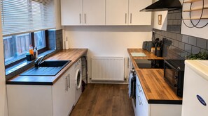 A compact kitchen with white cabinets, a wooden countertop, a built-in washing machine, and a black stove. - Creswell 2-bed House, Sleeps 7 With Street Parking (Worksop)