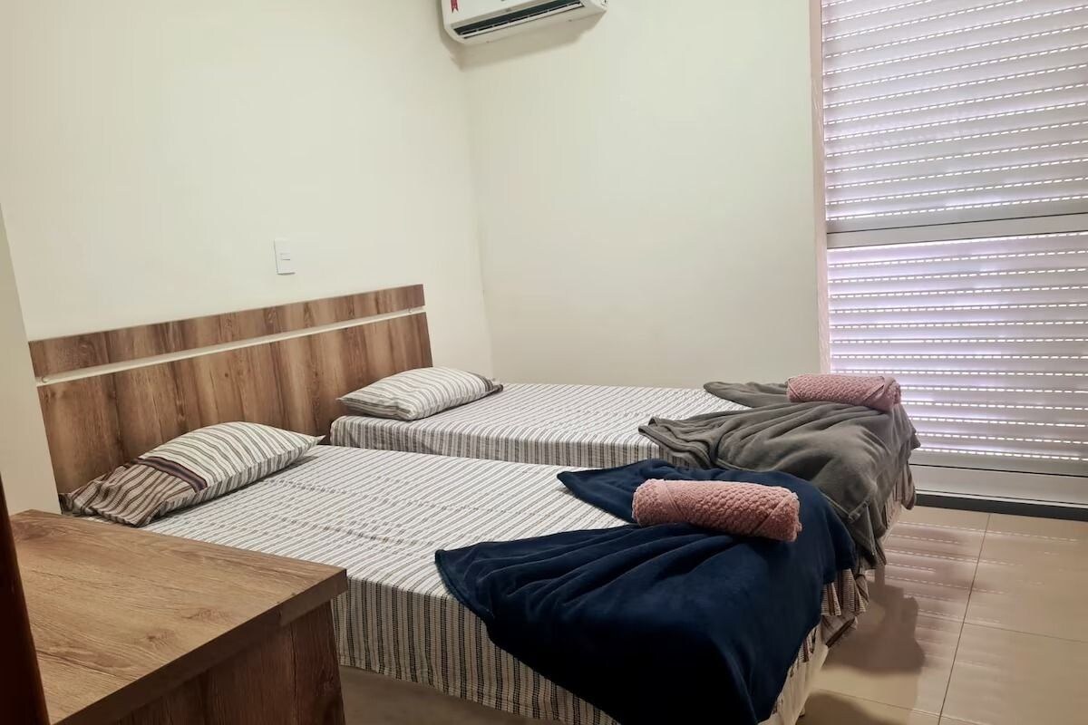 A bedroom with two beds, a wooden headboard, a bedside table, and a window with blinds.