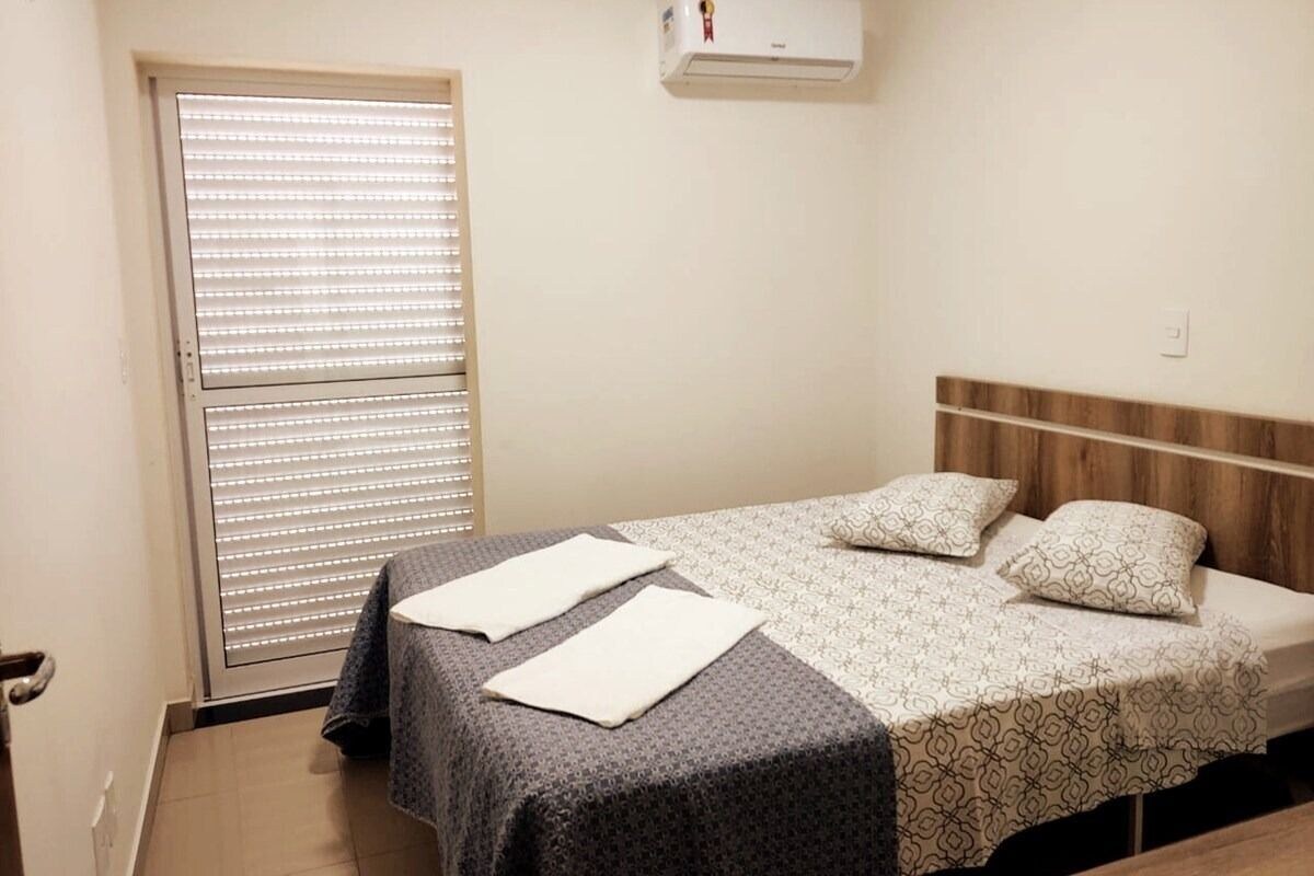 A bedroom with a bed, a window with blinds, a wall-mounted air conditioner, and a door.