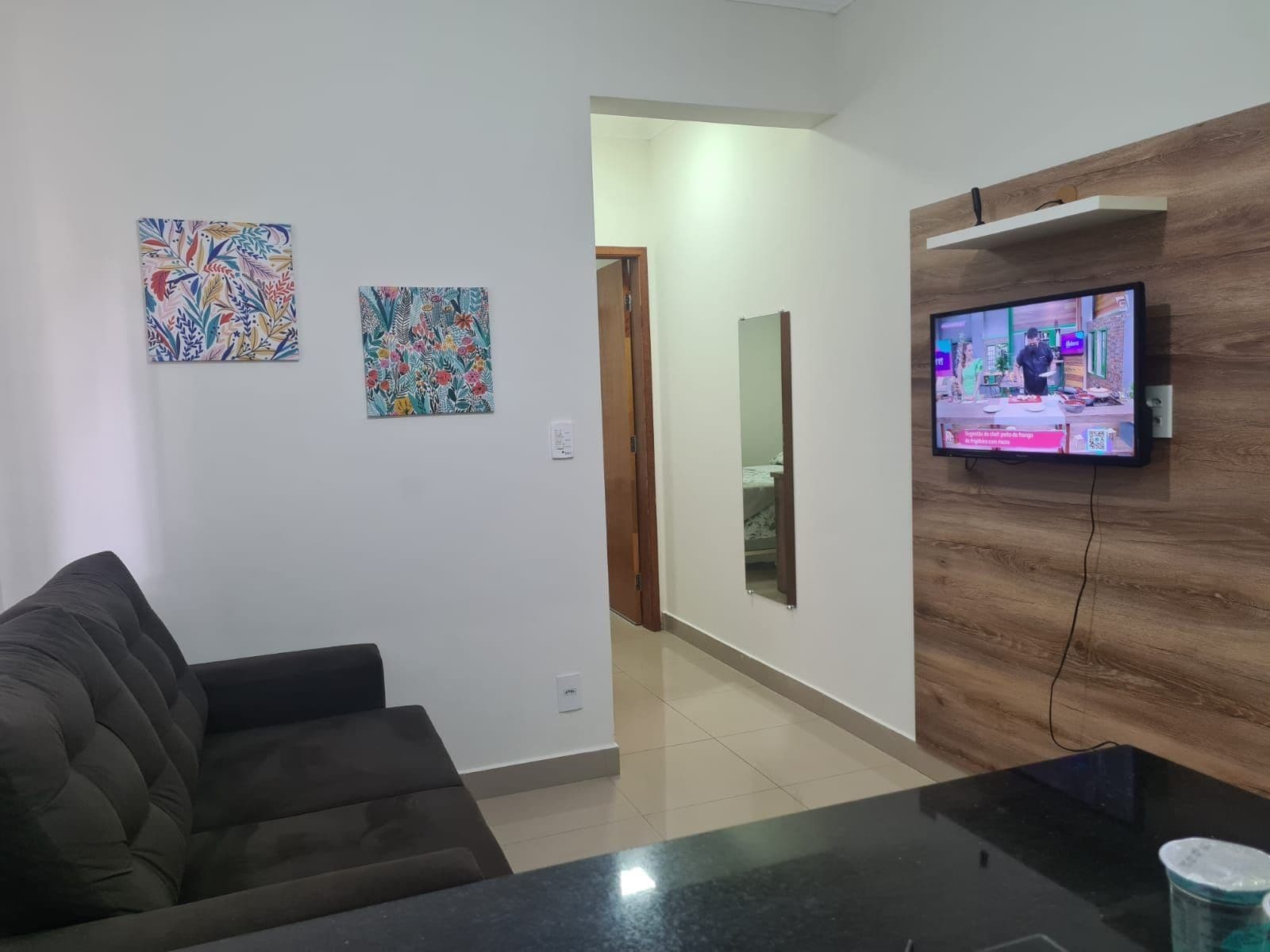 Sala de estar com um sofá escuro, duas obras de arte coloridas na parede, uma televisão em uma parede de madeira e uma mesa de centro com tampo de vidro.