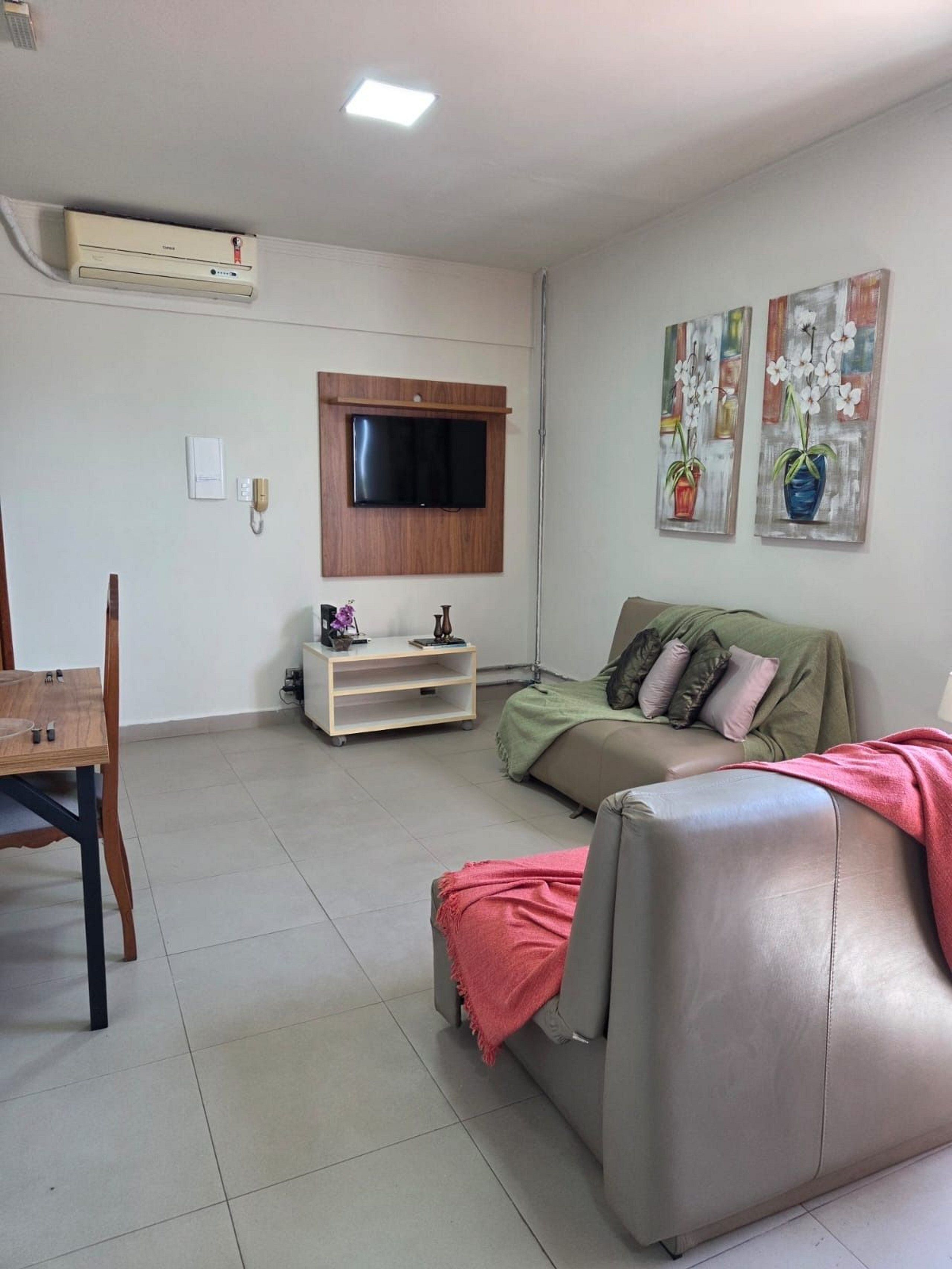 Uma sala de estar com televisão de tela plana na parede, um sofá, uma mesa de centro e uma pequena mesa de madeira com uma cadeira.