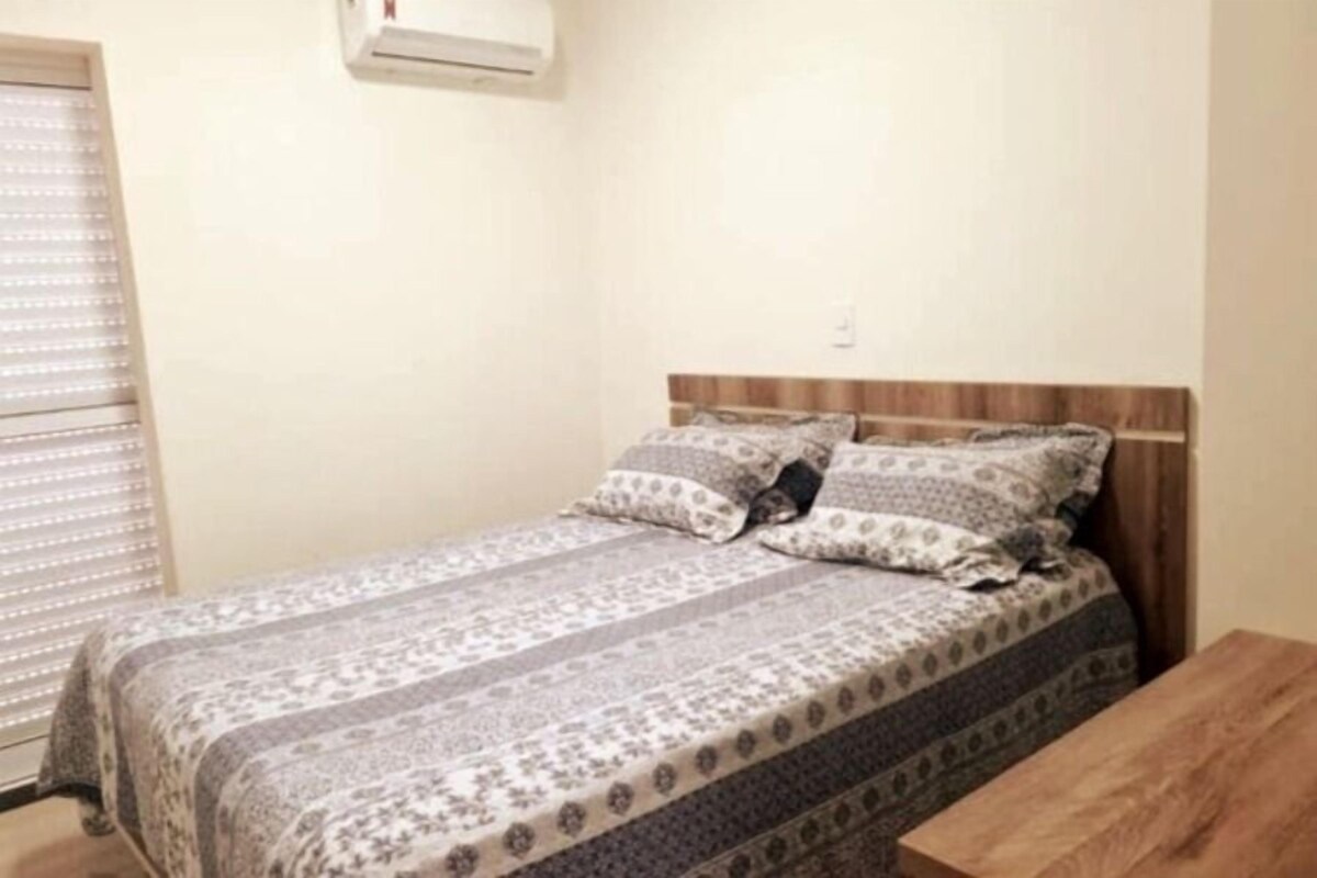 A single bed with a patterned bedspread and pillows, a wooden headboard, a small wooden table, and a window with blinds.