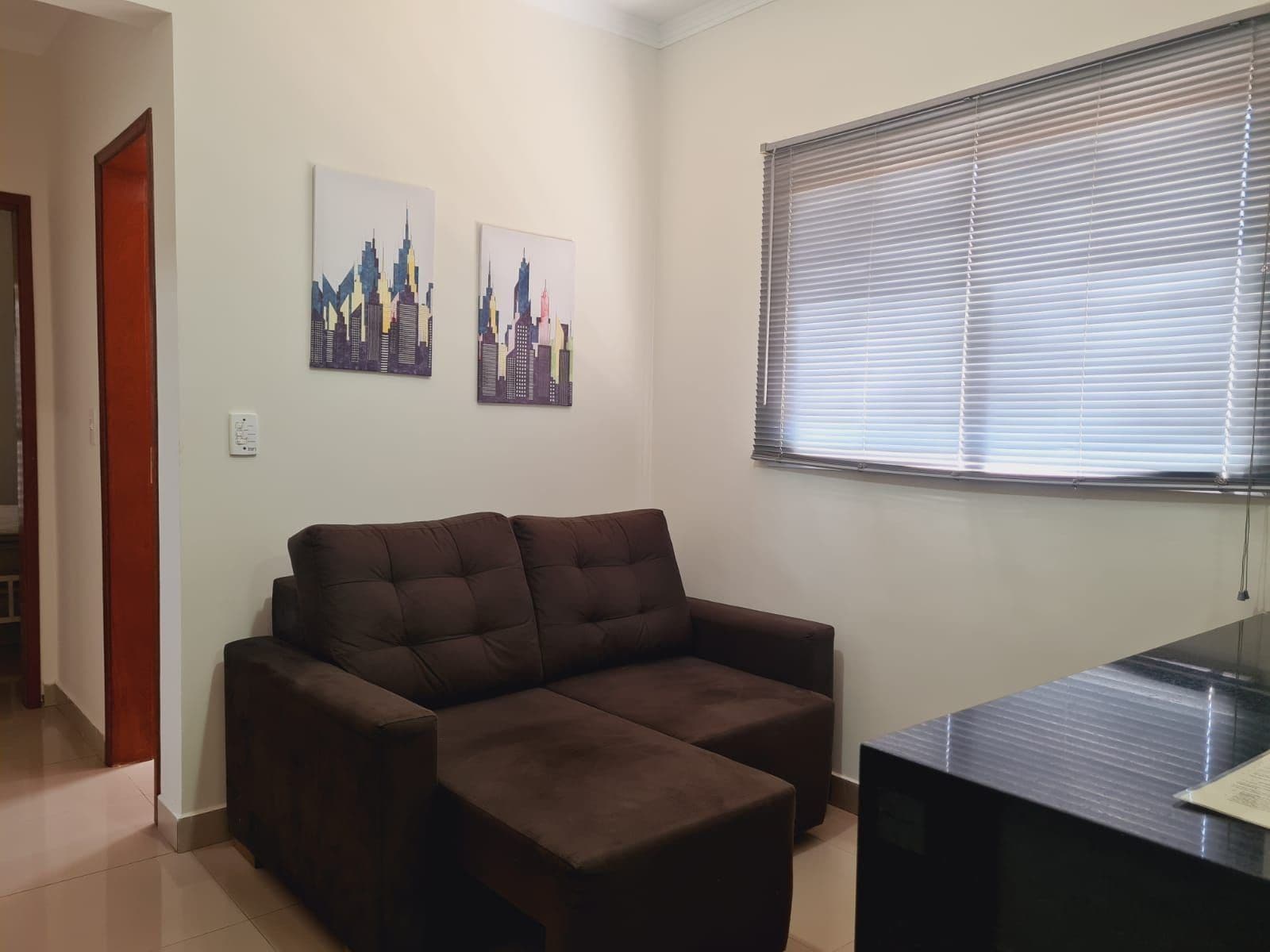 A living room with a brown sofa, two cityscape paintings on the wall, a window with blinds, and a black TV stand.