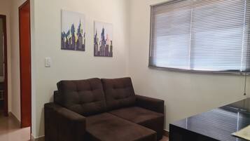 A living room with a brown sofa, two cityscape paintings on the wall, a window with blinds, and a black TV stand.