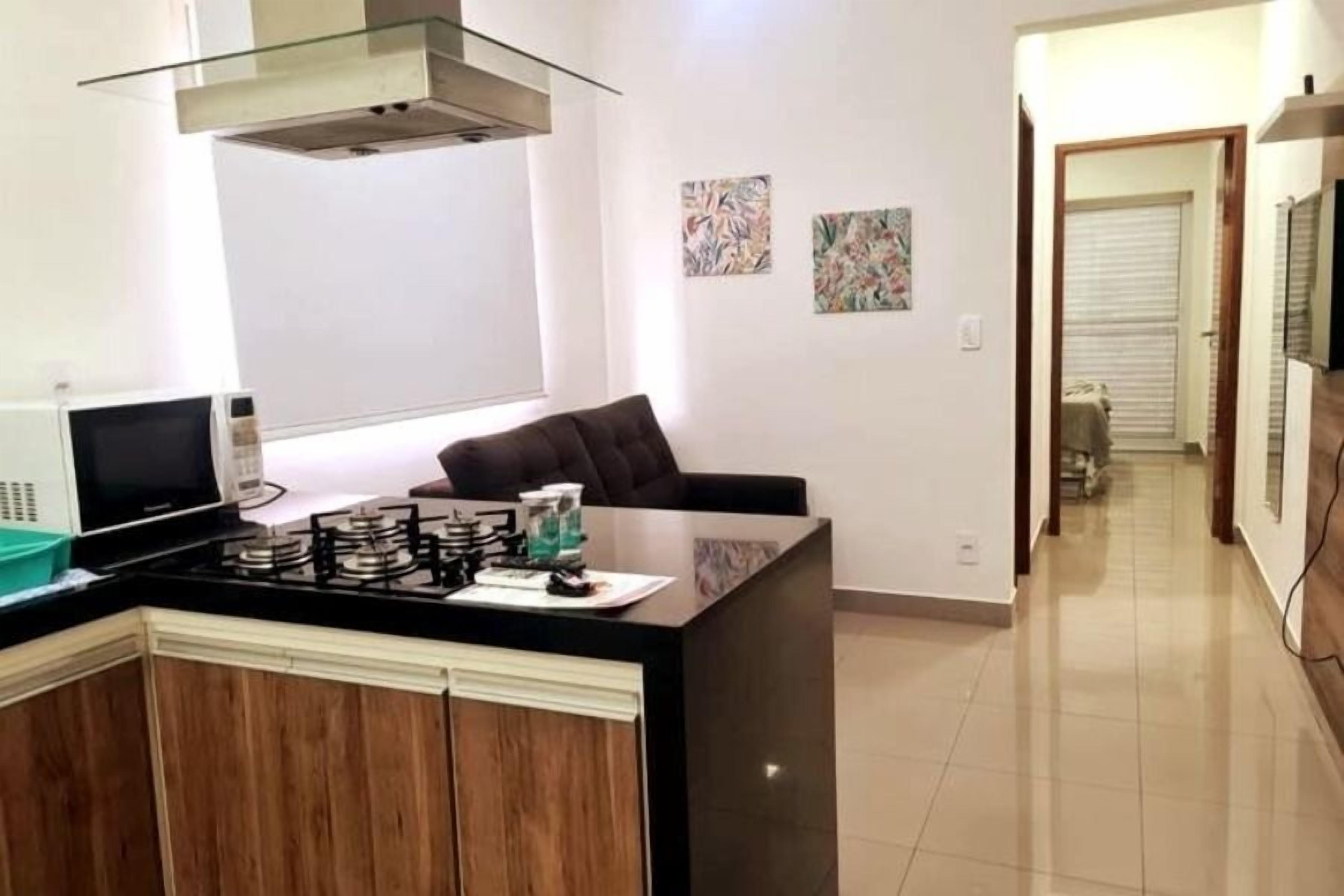 Cozinha moderna com fogão a gás, micro-ondas e máquina de café. Possui área de estar com sofá e poltrona. A cozinha tem armários de madeira e bancada preta.