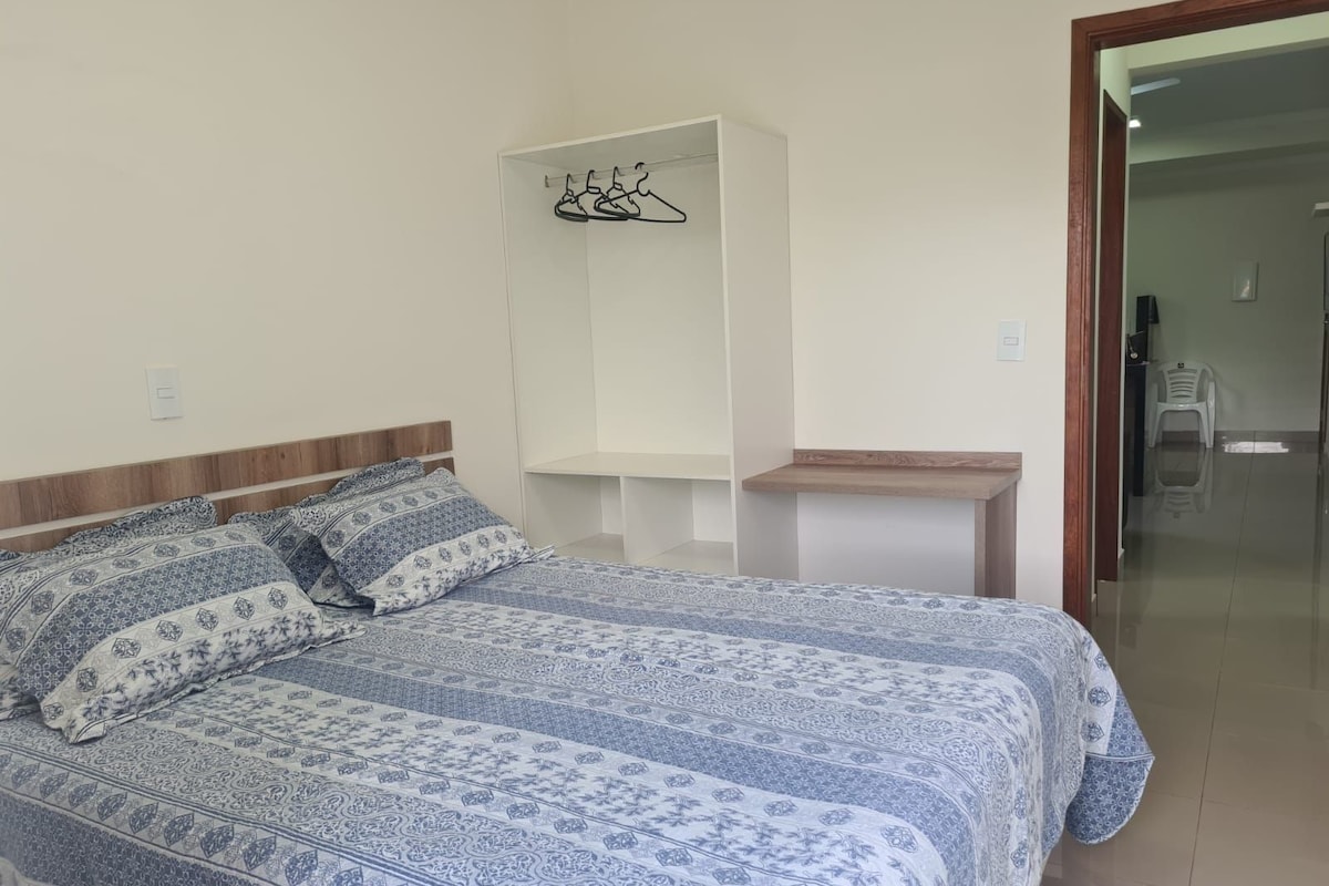 A bedroom with a bed, a bedside table, a hanging shelf, and a door leading to another room.