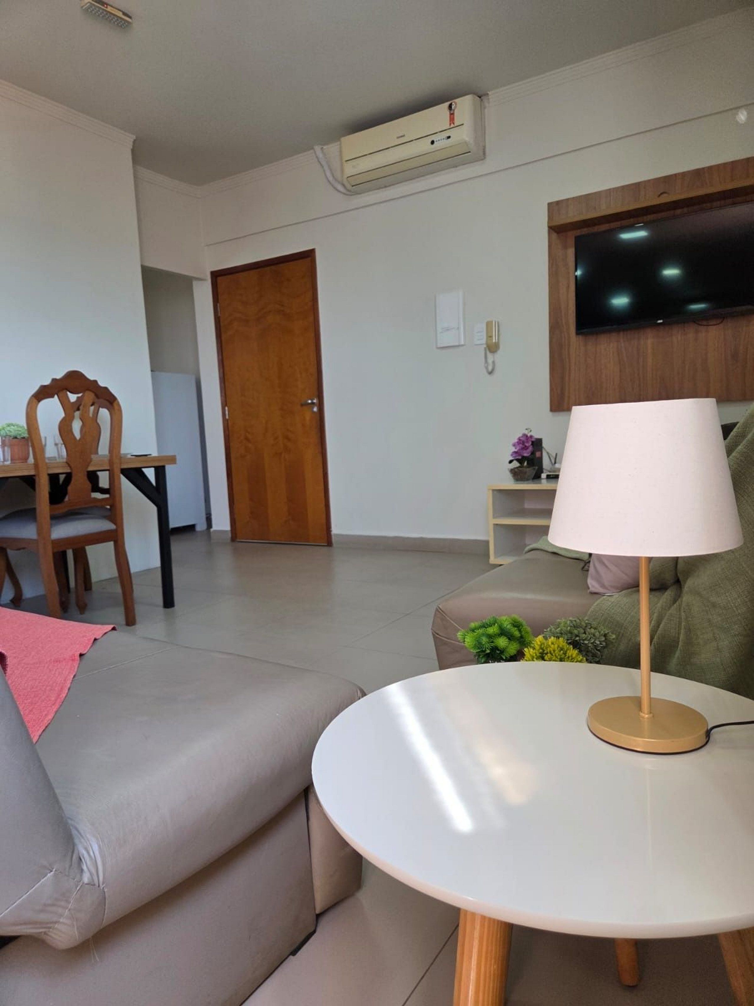 Uma sala de estar moderna com um sofá, uma mesa redonda com abajur, uma TV fixada na parede e uma porta de madeira.