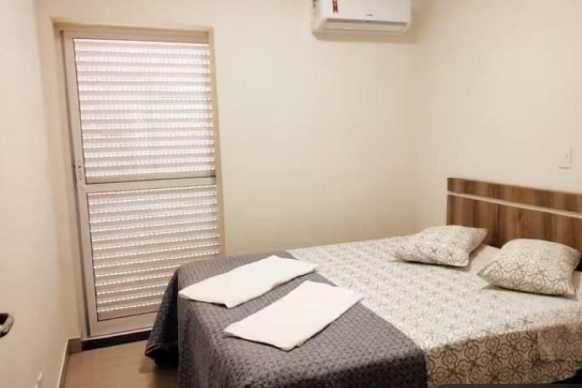 A hotel room with a bed, a window with blinds, and an air conditioning unit on the wall.