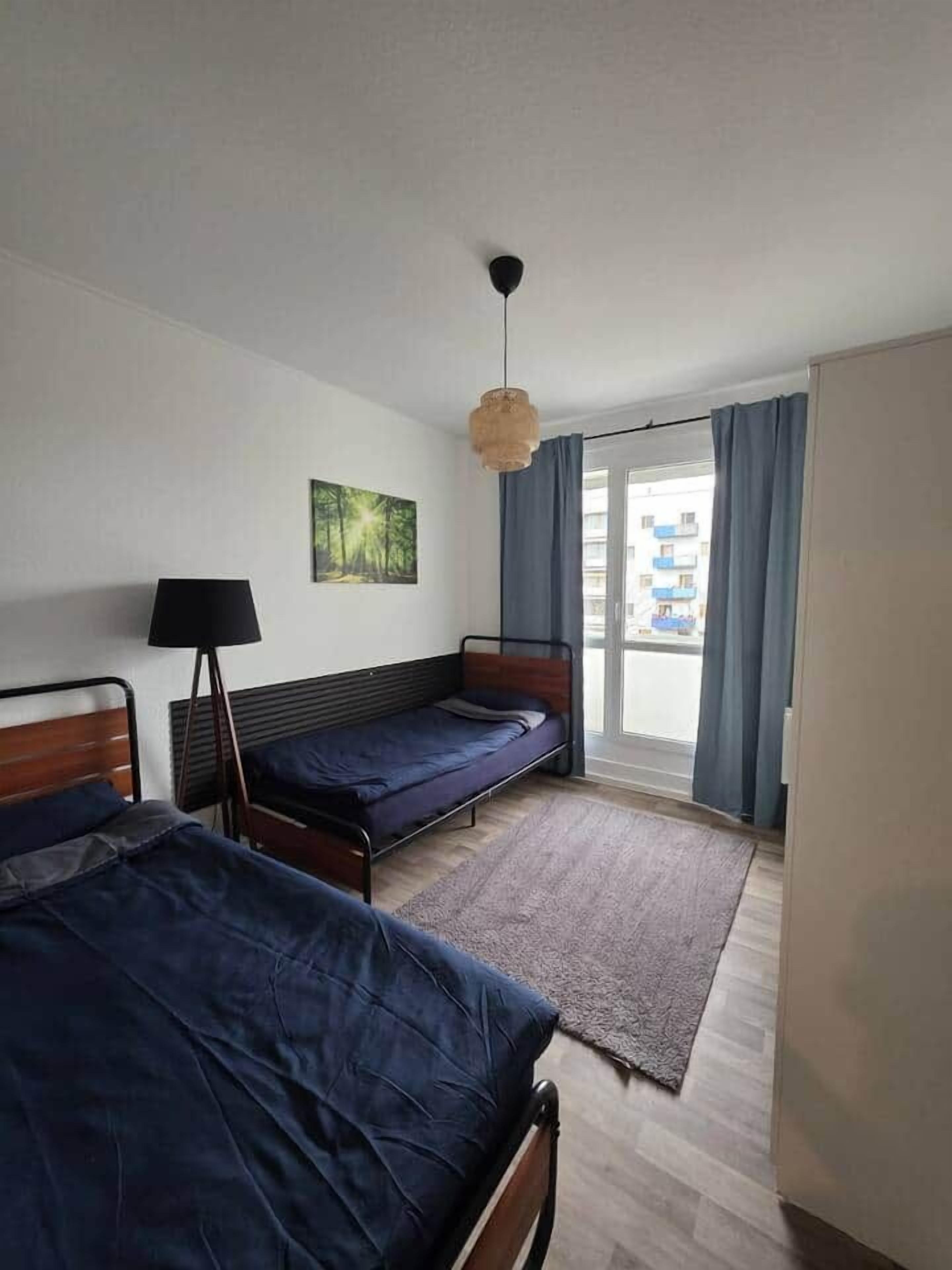 Ein Zimmer mit zwei Einzelbetten, einer Stehlampe, einem Fenster mit Vorhängen und Blick auf umliegende Gebäude.
